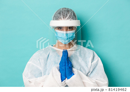 Concept of covid-19, pandemic and quarantine. Close-up of hopeful female doctor in personal protective equipment, holding hands in pray, begging or say please, standing over blue background Concept of covid-19, pandemic and quarantine. Close-up of hopeful female doctor in personal protective equipment, holding hands in pray, begging or say please, standing over blue background 81419462
