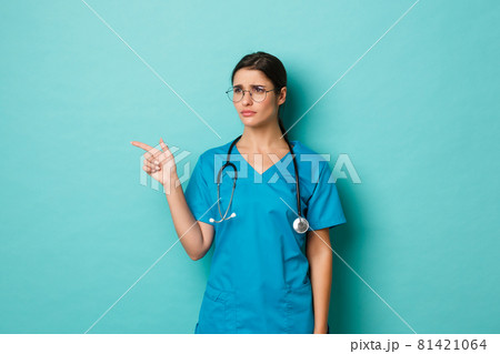Coronavirus, pandemic and social distancing concept. Image of beautiful sad female doctor, wearing scrubs and glasses, frowning upset, pointing fingers left at copy space, blue background Coronavirus, pandemic and social distancing concept. Image of beautiful sad female doctor, wearing scrubs and glasses, frowning upset, pointing fingers left at copy space, blue background 81421064