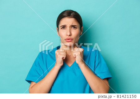 Coronavirus, social distancing and health concept. Close-up of worried young nurse in scrubs, looking concerned at something bad, grimacing disturbed, standing over blue background Coronavirus, social distancing and health concept. Close-up of worried young nurse in scrubs, looking concerned at something bad, grimacing disturbed, standing over blue background 81421150