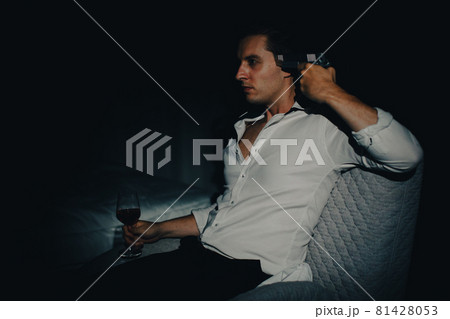 A young man in casual business clothes sitting alone in dark bedroom holding a pistol aim at his head and wine glass looks like going to kill himself. Concept of hopelessness and give up people 81428053