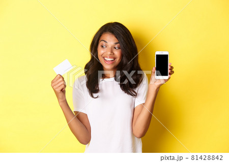 Image of excited african-american female model, showing smartphone screen and credit card, standing over yellow background 81428842