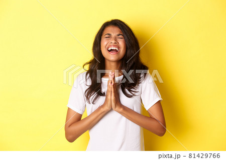 Portrait of young african-american girl pleading, begging for help with hands pressed together on chest, standing in white t-shirt over yellow background 81429766