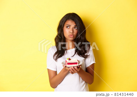 Image of indecisive and sad african-american woman, looking upset, cannot eat cake, standing over yellow background Image of indecisive and sad african-american woman, looking upset, cannot eat cake, standing over yellow background 81431440