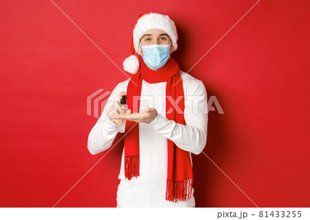 Concept of covid-19, christmas and holidays during pandemic. Happy young man in santa hat and medical mask, sanitize hands with antiseptic and smiling, standing over red background Concept of covid-19, christmas and holidays during pandemic. Happy young man in santa hat and medical mask, sanitize hands with antiseptic and smiling, standing over red background 81433255