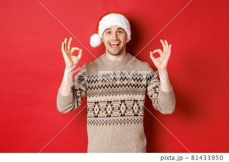 Portrait of happy and pleased handsome man, wearing winter sweater and santa hat, showing okay signs, praise good christmas party, standing over red background 81433950