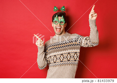 Portrait of joyful handsome guy in party glasses and christmas sweater, dancing and pointing fingers sideways, enjoying new year celebration, standing over red background 81433979