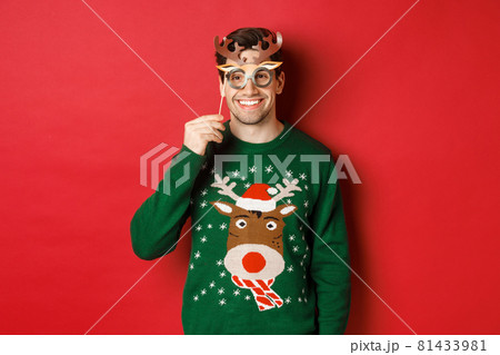 Handsome happy man in christmas sweater, holding party mask and smiling, enjoying new year celebration, standing over red background 81433981