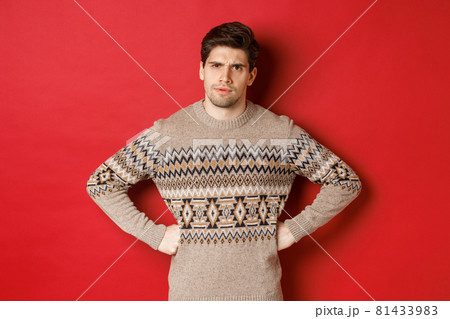 Portrait of doubtful and disappointed man, wearing christmas sweater, frowning and looking displeased at you, standing against red background 81433983