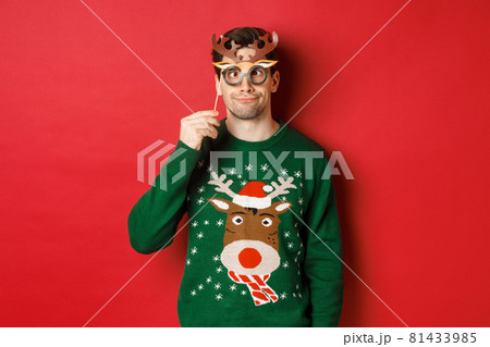 Funny man in christmas sweater and party mask, celebrating winter holidays, showing funny faces, standing against red background 81433985