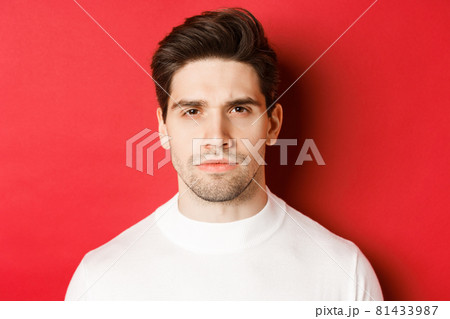 Close-up of thoughtful, serious-looking man in white sweater, standing over red background 81433987
