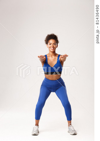 Full length of attractive african-american fit woman in sport clothing, stretch hands foward and making squats exercises, smiling at camera, standing against white background 81436490