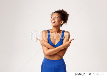 Attractive african-american fitness woman in blue sportswear, pointing fingers sideways and looking at upper left corner, looking happy, standing against white background Attractive african-american fitness woman in blue sportswear, pointing fingers sideways and looking at upper left corner, looking happy, standing against white background 81436692