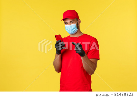 Food delivery, tracking orders, covid-19 and self-quarantine concept. Young courier, employee in medical mask and gloves, using mobile phone and pointing finger camera, yellow background Food delivery, tracking orders, covid-19 and self-quarantine concept. Young courier, employee in medical mask and gloves, using mobile phone and pointing finger camera, yellow background 81439322