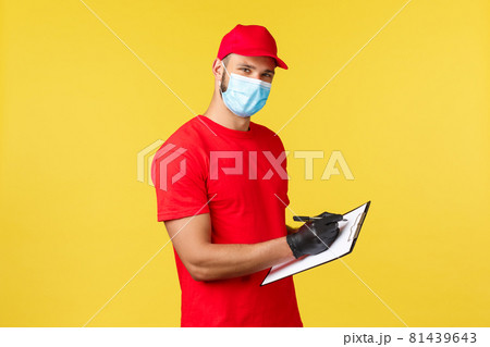 Express delivery during pandemic, covid-19, safe shipping, online shopping concept. Pleasant young courier in red uniform and medical mask, fill in order on clipboard, look camera 81439643