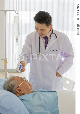 handsome asian male doctor wearing white lab coat with stethoscope stand checking temperature from old fat asian patient with gray hair wearing eyeglasses lay down on hospital bed with thermometer in 81443705