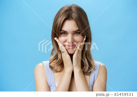 Pretty european girl pressing palms to cheeks and smiling happily while looking at camera as if checking out appereance in mirror liking result after applying skincare treatment mask over blue wall 81459531