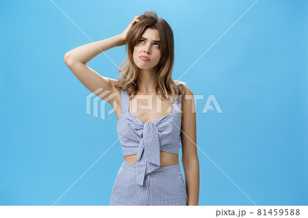 Silly confused charming adult female with chestnut hair in trendy outfit sulking looking questioned up with smirk and gloomy expression thinking scarthing head clueless over blue background 81459588