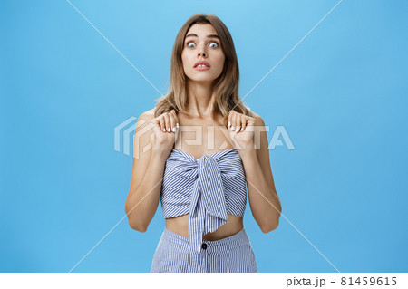 Portrait of stunned speechless and concerned young glamorous female pressing hands to chest popping eyes at camera standing in intense awkward and frightened pose being in stupor against blue wall 81459615