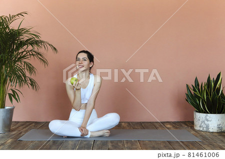 Young fit sporty woman in white sportswear sitting in lotus pose on yoga mat and eating green apple 81461006