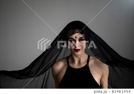 Redhead woman with a black veil on her head in a dark studio. A girl with an earring in her nose smiles slyly. Witch Makeup. Halloween costume. 81461454