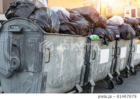 Rows of many big metal overloaded dumpster cans full of black plastic trash litter bags near residential building at city downtown or suburban area. Non-recyclable sorting garbage collecting 81467679