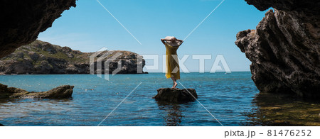 A woman in a white dress and a black hat standing on the rocks by the sea 81476252