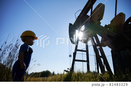 Side low view of child boy who standing opposite metal fence and examining oil pump structure in summer sunny day. On the background cloudless blue sky. 81483329