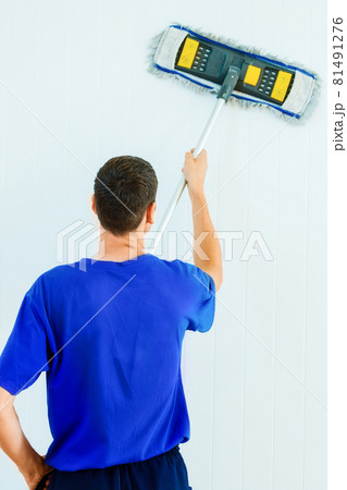 A young Caucasian man in a blue T-shirt washes the white wall of the room with a mop. A young Caucasian man in a blue T-shirt washes the white wall of the room with a mop. 81491276