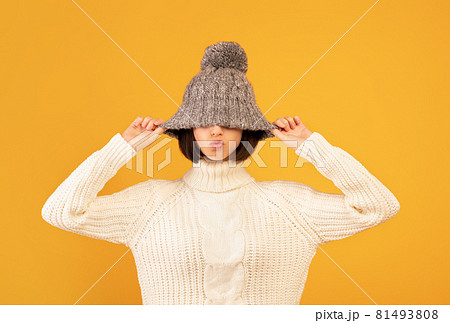 Pretty young woman pulling down woollen hat, showing kissing lips, fooling and posing to camera over yellow background 81493808