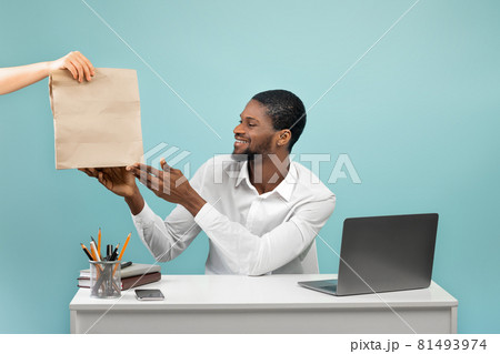 Millennial black businessman receiving food delivery from courier, sitting at workplace over blue studio background Millennial black businessman receiving food delivery from courier, sitting at workplace over blue studio background 81493974