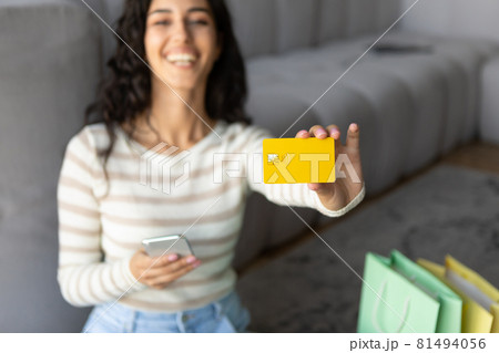 Happy young woman showing credit card at camera, holding smartphone, shopping in online store from home, selective focus Happy young woman showing credit card at camera, holding smartphone, shopping in online store from home, selective focus 81494056