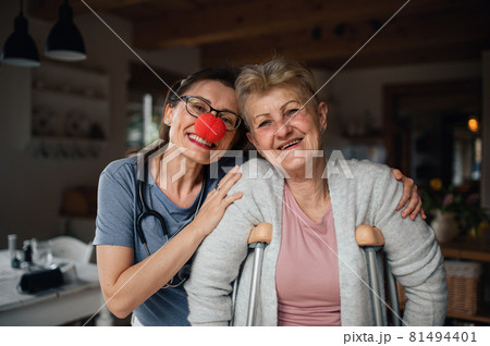 Healthcare worker or caregiver with red nose visiting senior woman indoors at home, looking at camera. 81494401