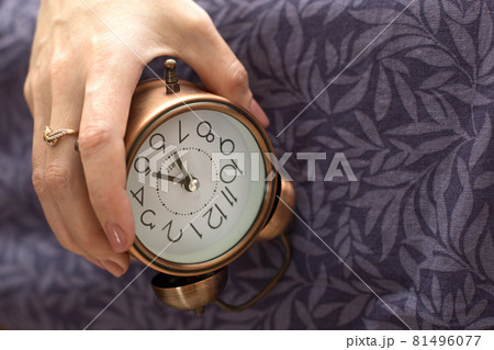 A woman's hand with an alarm clock hanging from the bed. The concept of a difficult awakening, an early rise A woman's hand with an alarm clock hanging from the bed. The concept of a difficult awakening, an early rise 81496077