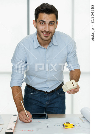 Young businessman standing in office beside working desk and working with home wooden model in hand Young businessman standing in office beside working desk and working with home wooden model in hand 81504268