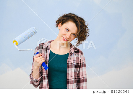 A young beautiful woman painter designer paints a white wall with blue paint with roller. Plaid shirt, short hair and jeans. A young beautiful woman painter designer paints a white wall with blue paint with roller. Plaid shirt, short hair and jeans. 81520545
