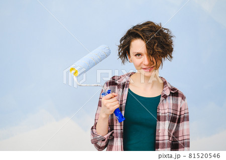 A young beautiful woman painter designer paints a white wall with blue paint with roller. Plaid shirt, short hair and jeans. 81520546