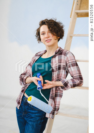 A young beautiful woman painter designer paints a white wall with blue paint with roller. Plaid shirt, short hair and jeans. 81520556