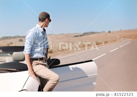 Handsome young man in sunglasses leaning on luxury white convertible while standing stop on road 81522525