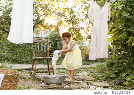 Little girl in dress washing clothes in metal basin while helping mom with chores in backyard 81522538