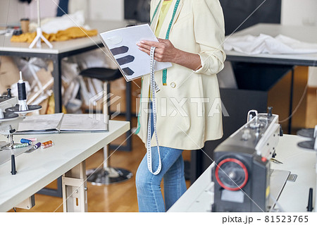 Middle aged seamstress holds tape measure and papers standing near tables with sewing machines Middle aged seamstress holds tape measure and papers standing near tables with sewing machines 81523765