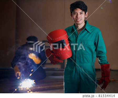 smart confident young Asian man wearing a factory uniform holding a welding helmet posing smiling to a camera with soft background on his colleague working on welding 81534414