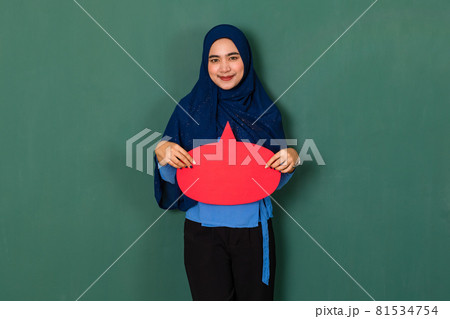 confident and happy woman concept. Pretty attractive Muslim lady dressed beautifully wearing blue hijab standing and holding a red blank speech bubble over green background confident and happy woman concept. Pretty attractive Muslim lady dressed beautifully wearing blue hijab standing and holding a red blank speech bubble over green background 81534754