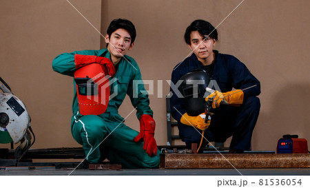 young handsome Asian mechanic sitting and smiling at a camera in a factory's workshop. They are wearing safety uniforms for welding work. Safety working concept 81536054