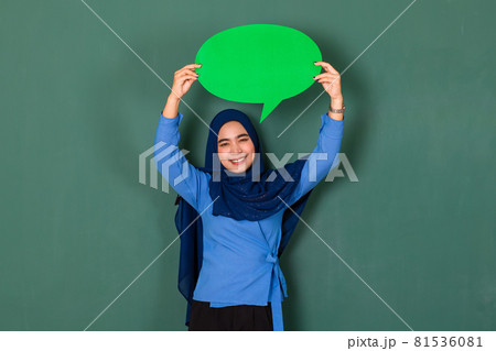 confident and happy woman concept. Pretty attractive Muslim lady dressed beautifully wearing blue hijab standing and holding green blank speech bubble over green background confident and happy woman concept. Pretty attractive Muslim lady dressed beautifully wearing blue hijab standing and holding green blank speech bubble over green background 81536081