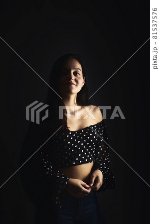 Half-length portrait of a young girl with a smile looking towards the camera, on a dark studio background Half-length portrait of a young girl with a smile looking towards the camera, on a dark studio background 81537576