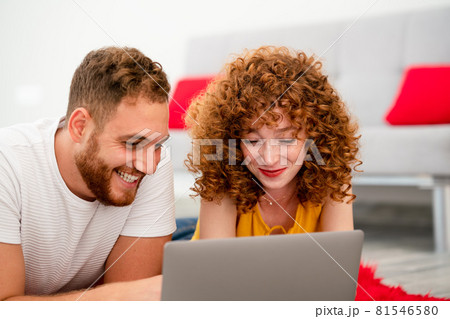 A Young couple lovers or friends laying on a red pluffy carpet and using laptop. 81546580