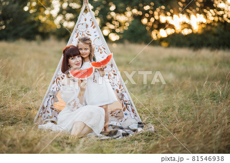 Mother and cute daughter dressed in boho style, sitting in front of tee-pee tent with watermelon 81546938