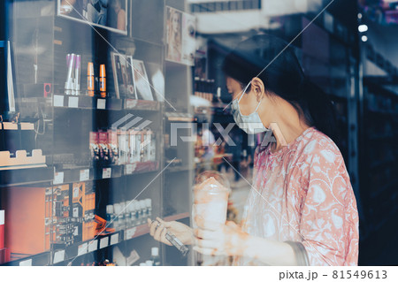 Woman in protective mask shopping in beauty and make up store shot through window. Woman in protective mask shopping in beauty and make up store shot through window. 81549613