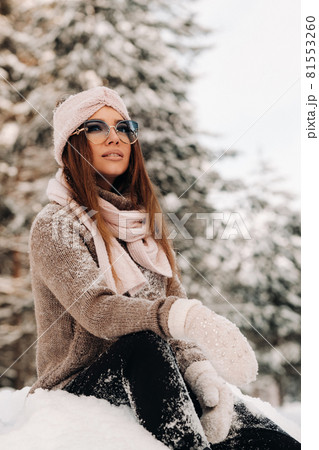 A girl in a sweater and glasses in winter sits on a snow-covered background in the forest 81553260