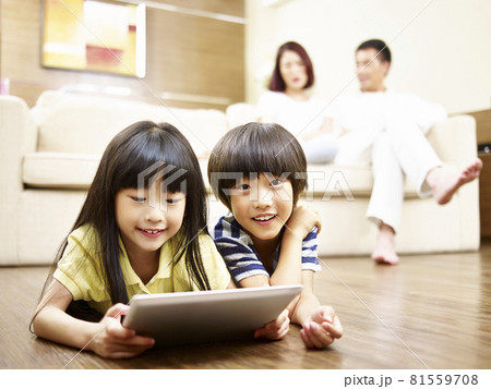 asian children lying on front on floor playing video game using ipad parents watching in background asian children lying on front on floor playing video game using ipad parents watching in background 81559708